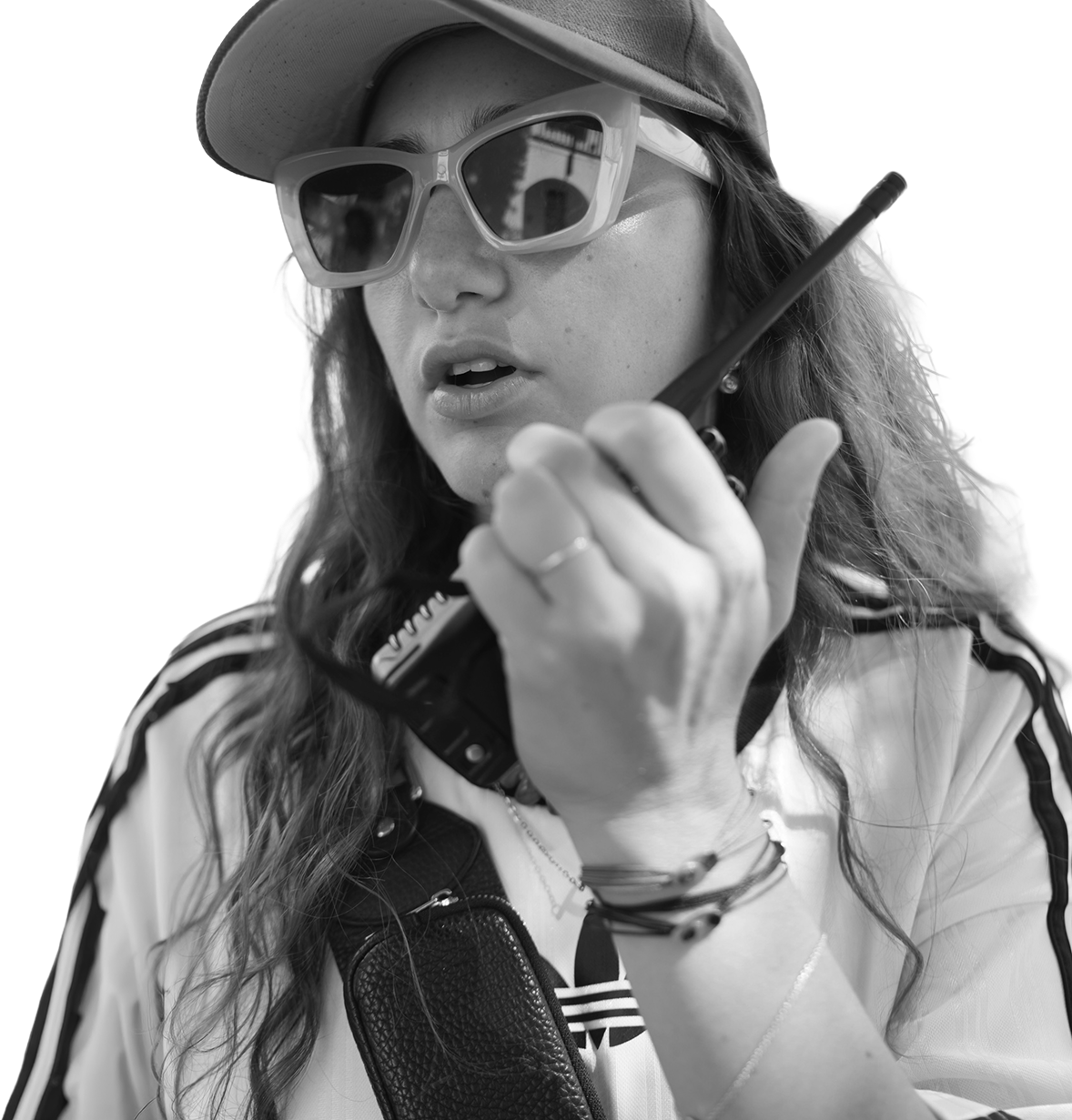 A woman wearing sunglasses and a cap speaks into a walkie-talkie. She appears focused and engaged, with long hair and a casual, sporty outfit.