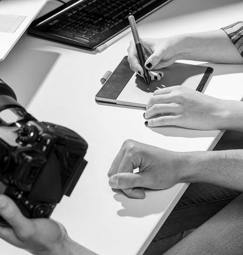 Two people work at a desk, one using a graphics tablet and pen, with a camera and keyboard nearby. The setting is creative and collaborative.