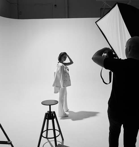 Black and white photo of a fashion photoshoot. A model stands confidently in a studio, lit by bright lights, while a photographer captures the moment.img