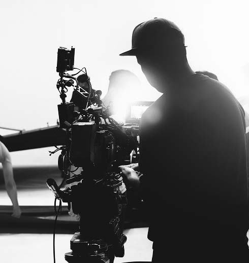 Silhouetted film crew operates a camera on a set, with bright light in the background. The scene conveys focus and creative intensity.