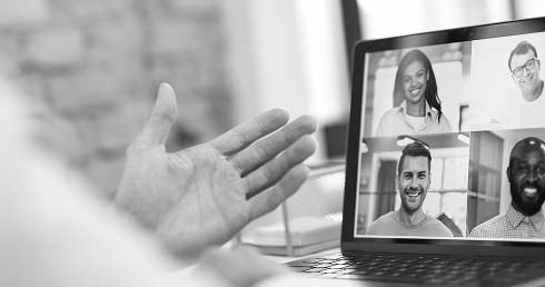 Black-and-white image of a laptop screen showing a video call with four smiling participants. A hand gestures in the foreground, suggesting interaction.