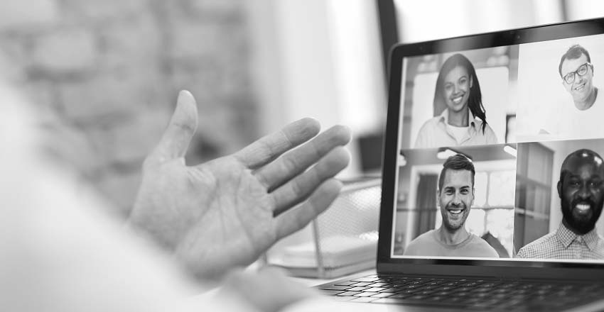 Black-and-white image of a laptop screen showing a video call with four smiling participants. A hand gestures in the foreground, suggesting interaction.