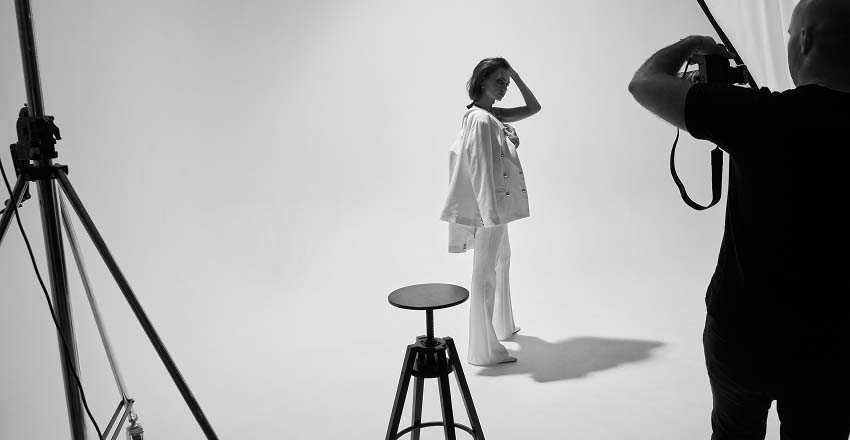Black and white photo of a fashion photoshoot. A model stands confidently in a studio, lit by bright lights, while a photographer captures the moment.