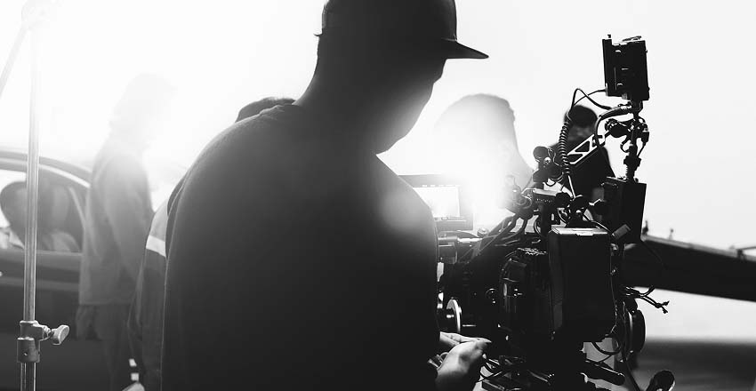Silhouetted film crew operates a camera on a set, with bright light in the background. The scene conveys focus and creative intensity.
