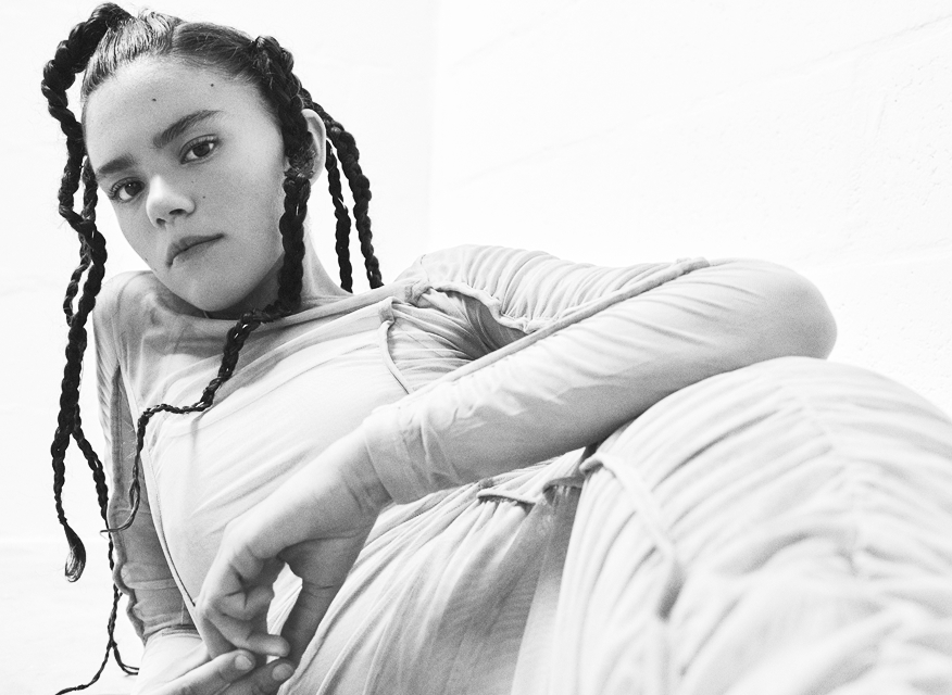 A black-and-white portrait of a person with braided hair, wearing a textured outfit. They recline against a white wall, exuding confidence and calmness.