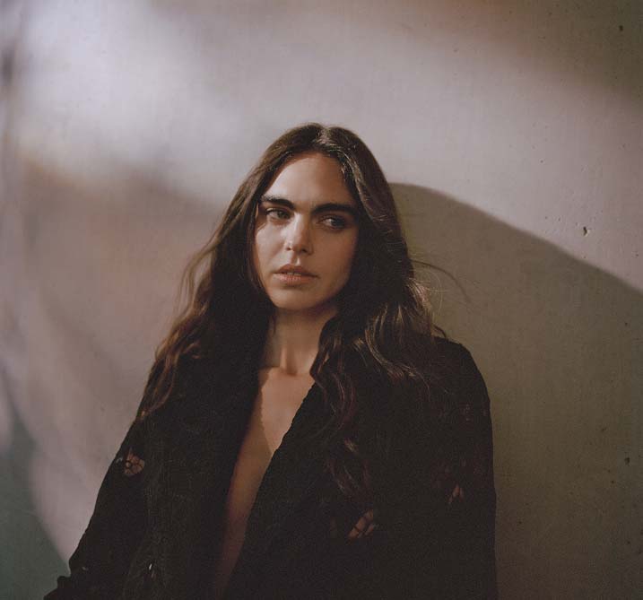 A woman with long dark hair leans against a shadowed wall, looking thoughtfully to the side. The dim lighting creates a contemplative and serene mood.