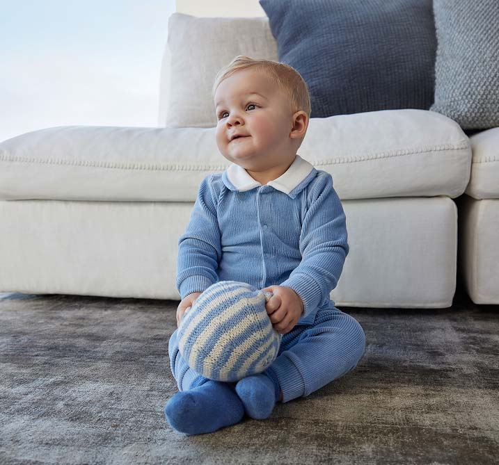 A baby sits on a textured rug, wearing a blue outfit, holding a striped blue and white ball. The scene is cozy with a soft sofa in the background.