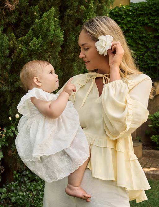 A woman in a pale yellow blouse stands in a garden, holding a baby in a white dress. They gaze at each other, exuding a loving and serene mood.