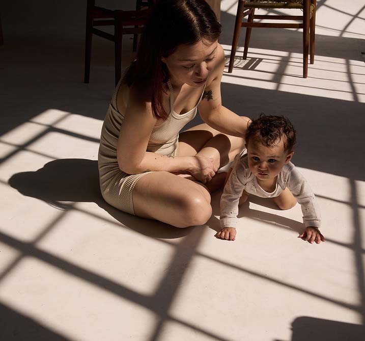 A woman in a beige outfit sits on the floor with a crawling baby in sunlight. She gently holds the baby, creating a warm, nurturing atmosphere. Shadows create patterns.