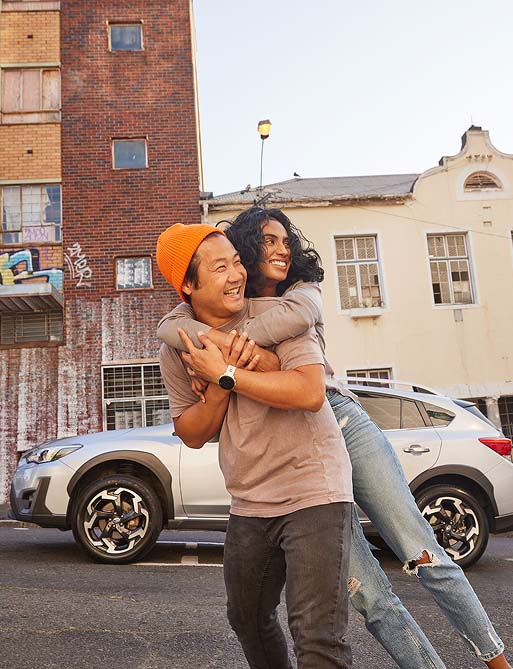 A joyful couple laughs as the man carries the woman piggyback on a street with colorful graffiti. A car and old buildings are in the background.