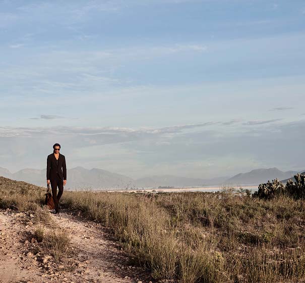 A person in a dark suit walks along a rocky path through a vast, grassy landscape under a clear blue sky, conveying a sense of solitude and tranquility.