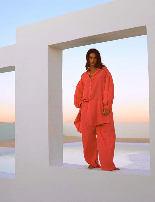 Person in oversized coral outfit stands in modern white architectural frame, with a serene sunset and pool in the background, exuding calmness.
