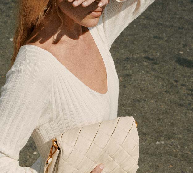 A person with long hair covers their face with a hand, wearing a white sweater. They are outdoors, casting a shadow on a sunlit, textured ground.
