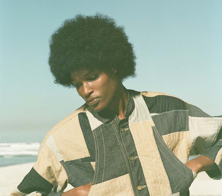 Person with an afro hairstyle wearing a patchwork jacket looks down thoughtfully against a serene beach background under a clear sky.