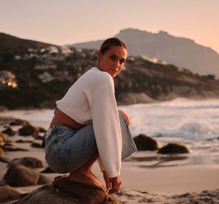 A person in a white sweater and jeans crouches on a rocky beach at sunset. The backdrop features mountains, waves, and an orange sky, conveying tranquility.