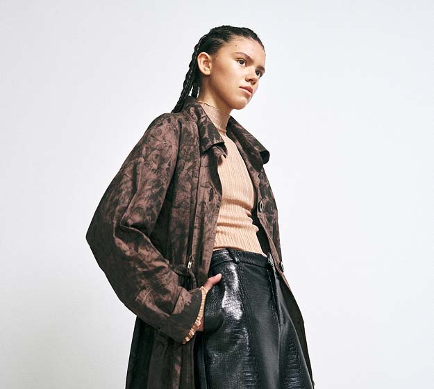 A model in a brown patterned coat, peach ribbed top, and black glossy pants stands confidently against a plain background, conveying a modern, edgy style.