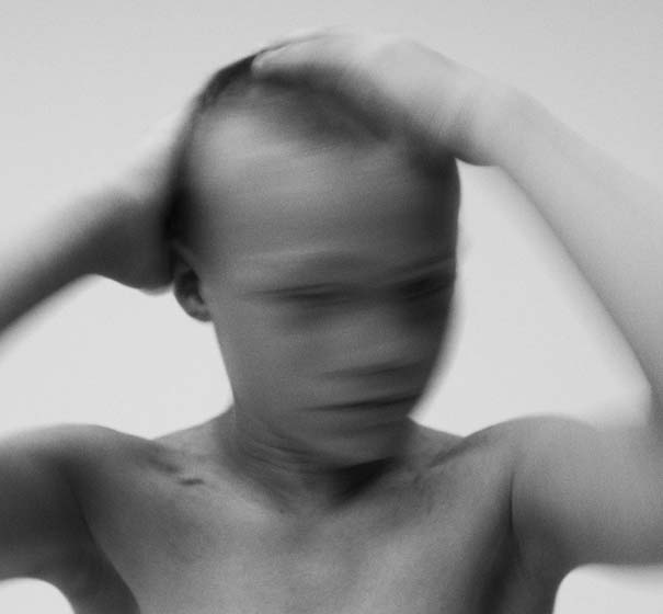 Blurred black and white image of a shirtless person with short hair, hands on head, conveying a sense of motion and introspection against a plain background.