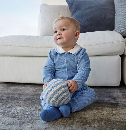 A baby sits on a textured rug, wearing a blue outfit, holding a striped blue and white ball. The scene is cozy with a soft sofa in the background.