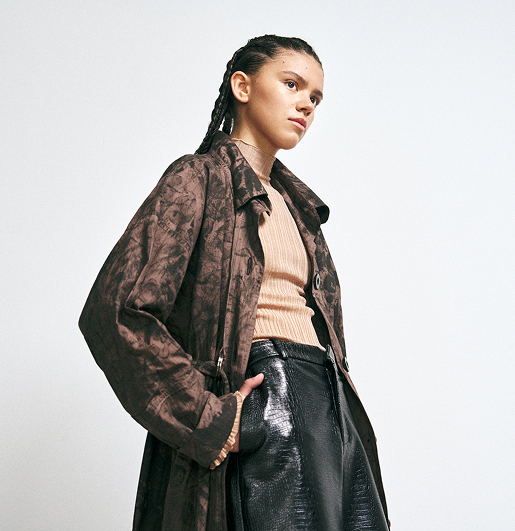 A model in a brown patterned coat, peach ribbed top, and black glossy pants stands confidently against a plain background, conveying a modern, edgy style.