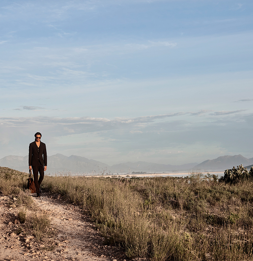 A person in a dark suit walks along a rocky path through a vast, grassy landscape under a clear blue sky, conveying a sense of solitude and tranquility.