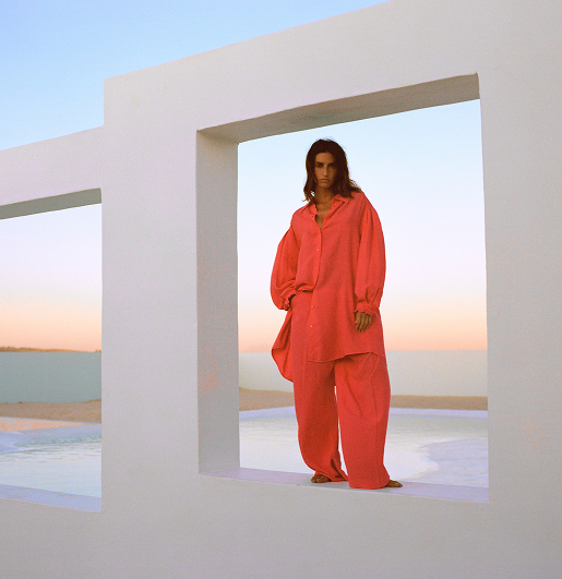 Person in oversized coral outfit stands in modern white architectural frame, with a serene sunset and pool in the background, exuding calmness.