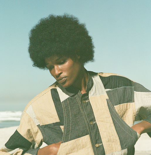 Person with an afro hairstyle wearing a patchwork jacket looks down thoughtfully against a serene beach background under a clear sky.