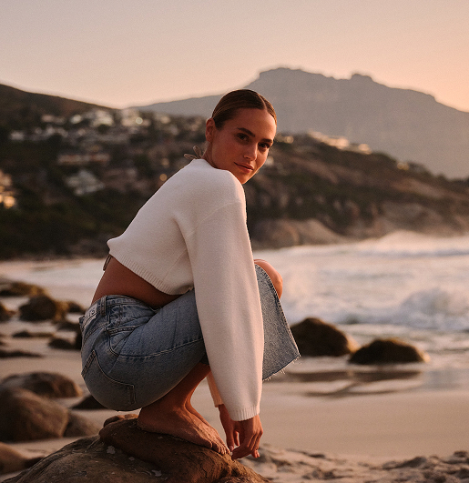 A person in a white sweater and jeans crouches on a rocky beach at sunset. The backdrop features mountains, waves, and an orange sky, conveying tranquility.